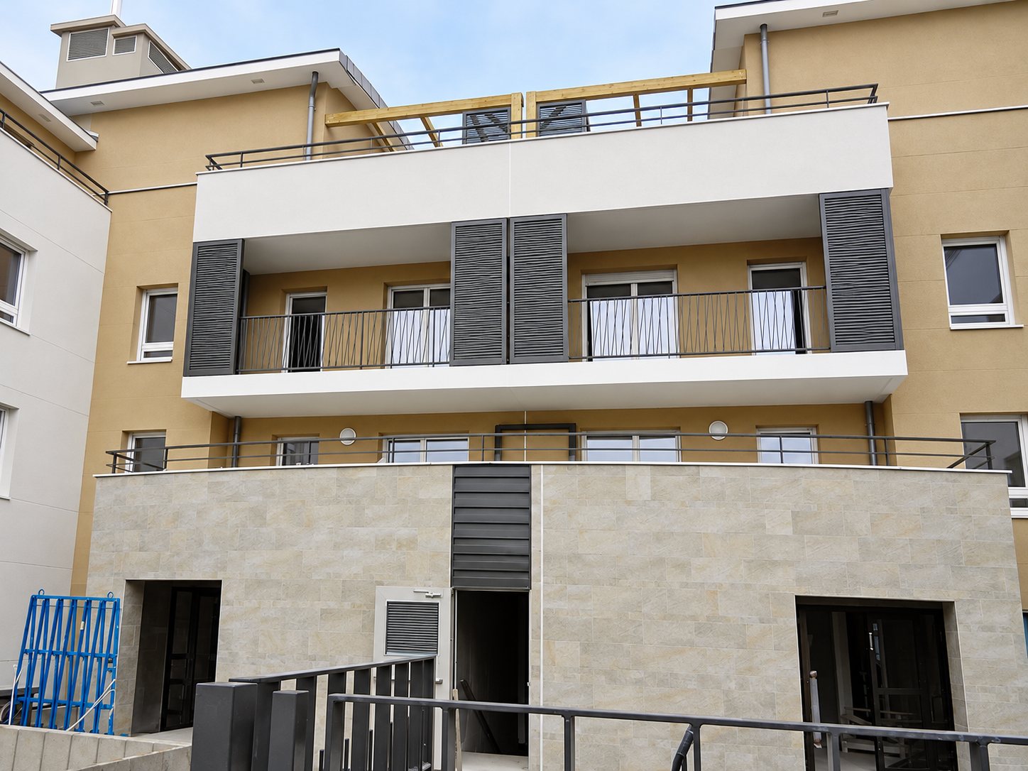 Façade beige à balcons et panneaux gris au-dessus d'un rez-de-chaussée en pierre claire.