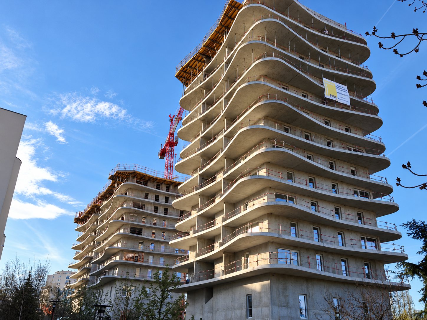 Immeuble en construction aux balcons arrondis, avec une grue et un autre bâtiment en arrière-plan sous un ciel bleu.