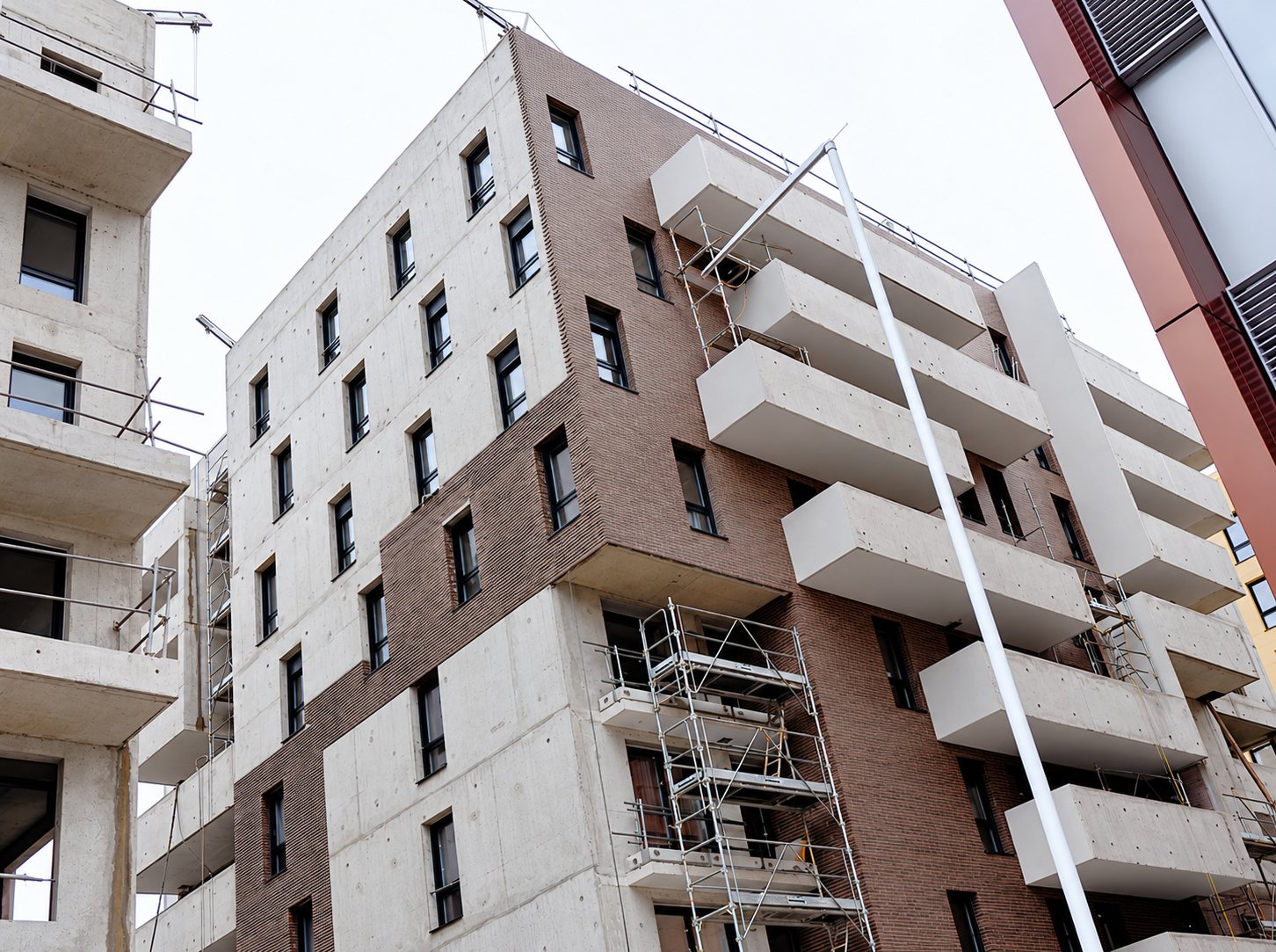 Vue en contre-plongée d'un immeuble résidentiel en construction, avec façades en béton et en brique, grands balcons blancs et échafaudages.