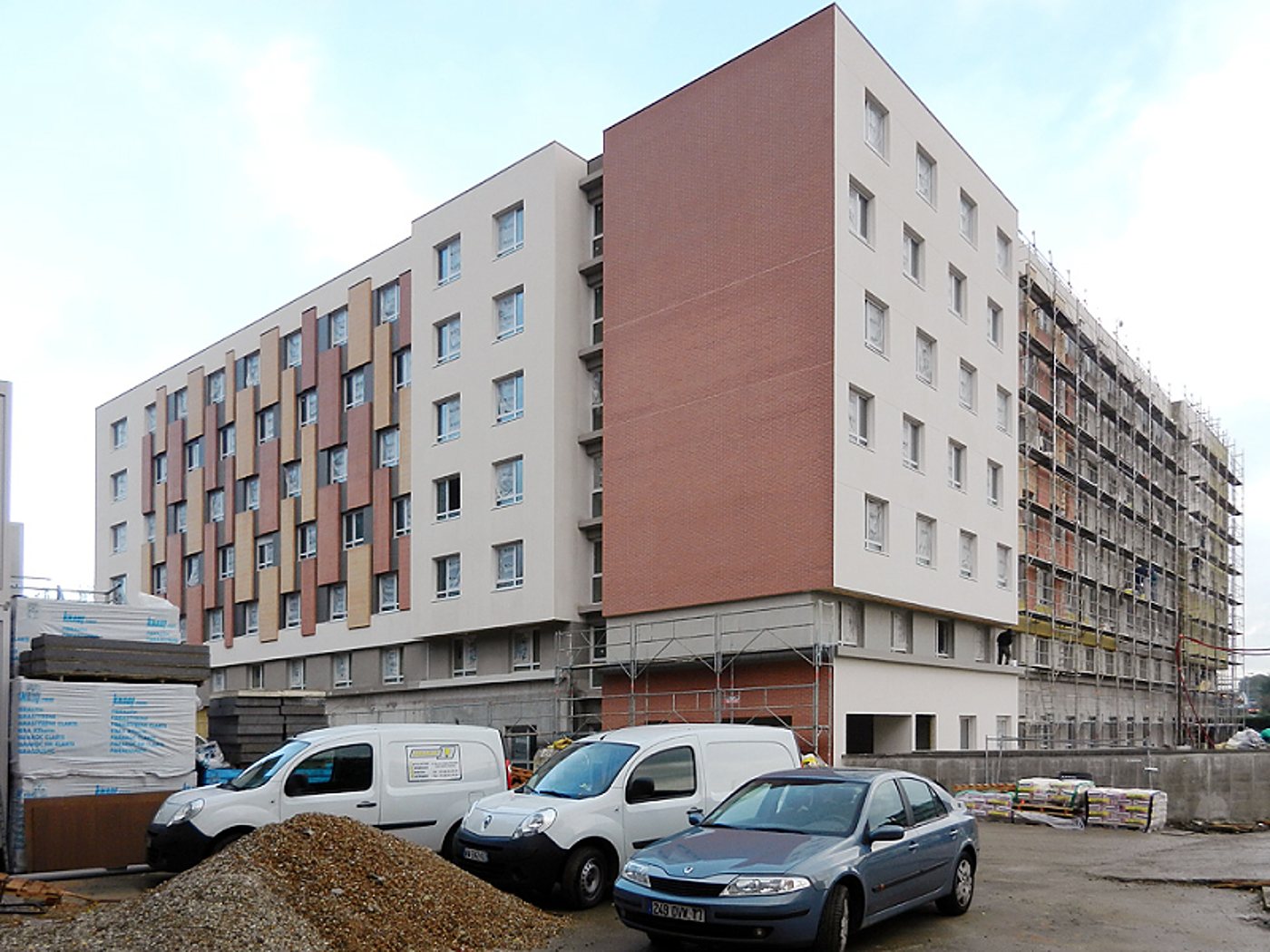 Un grand immeuble de logements aux façades blanches et rouges, en cours de finition avec des échafaudages sur le côté, et des camionnettes garées sur le chantier.