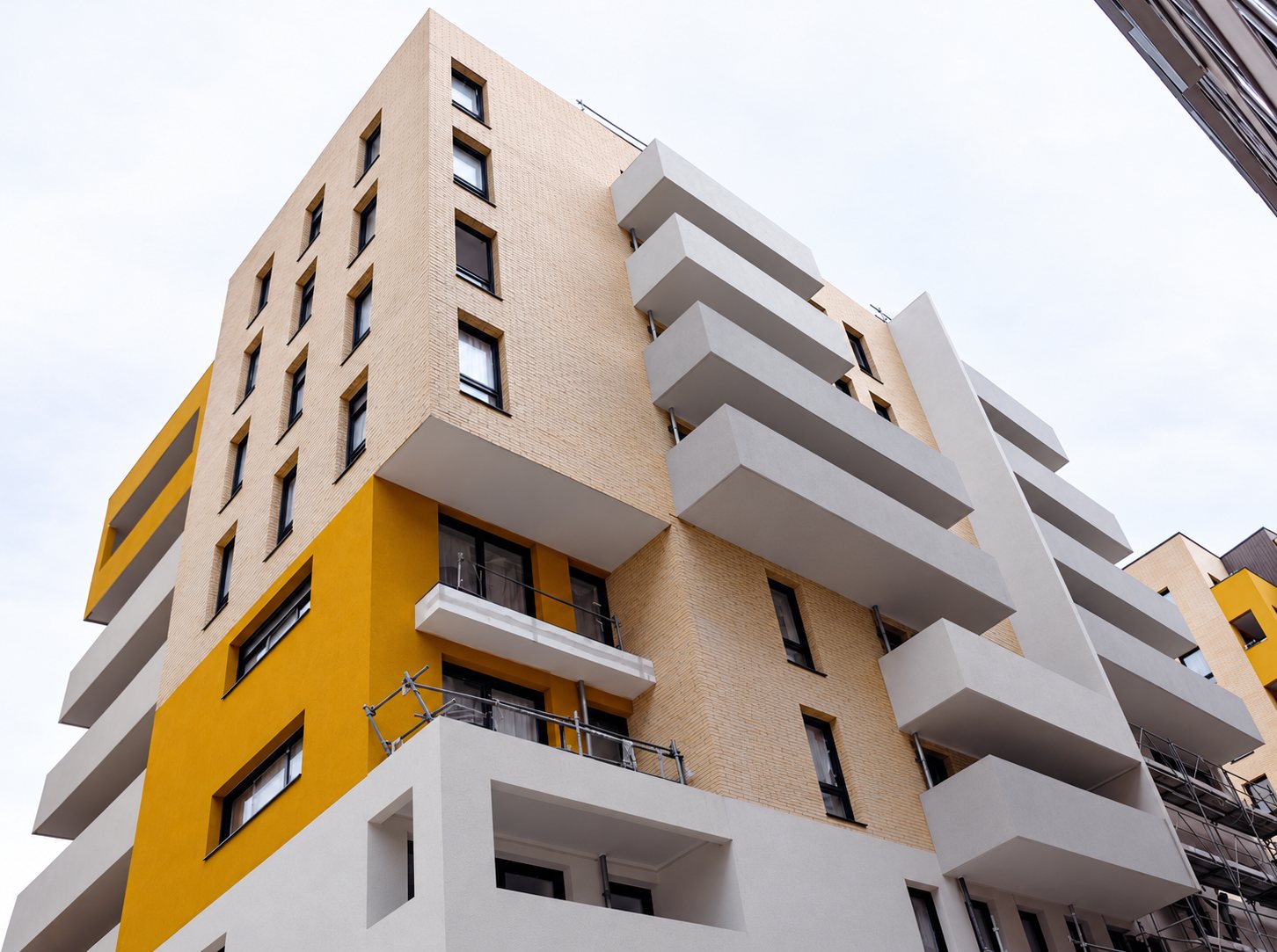 Vue en contre-plongée d'un immeuble d'habitation récent dont la façade combine briques claires, panneaux jaunes et larges balcons blancs, avec un échafaudage visible sur la droite.