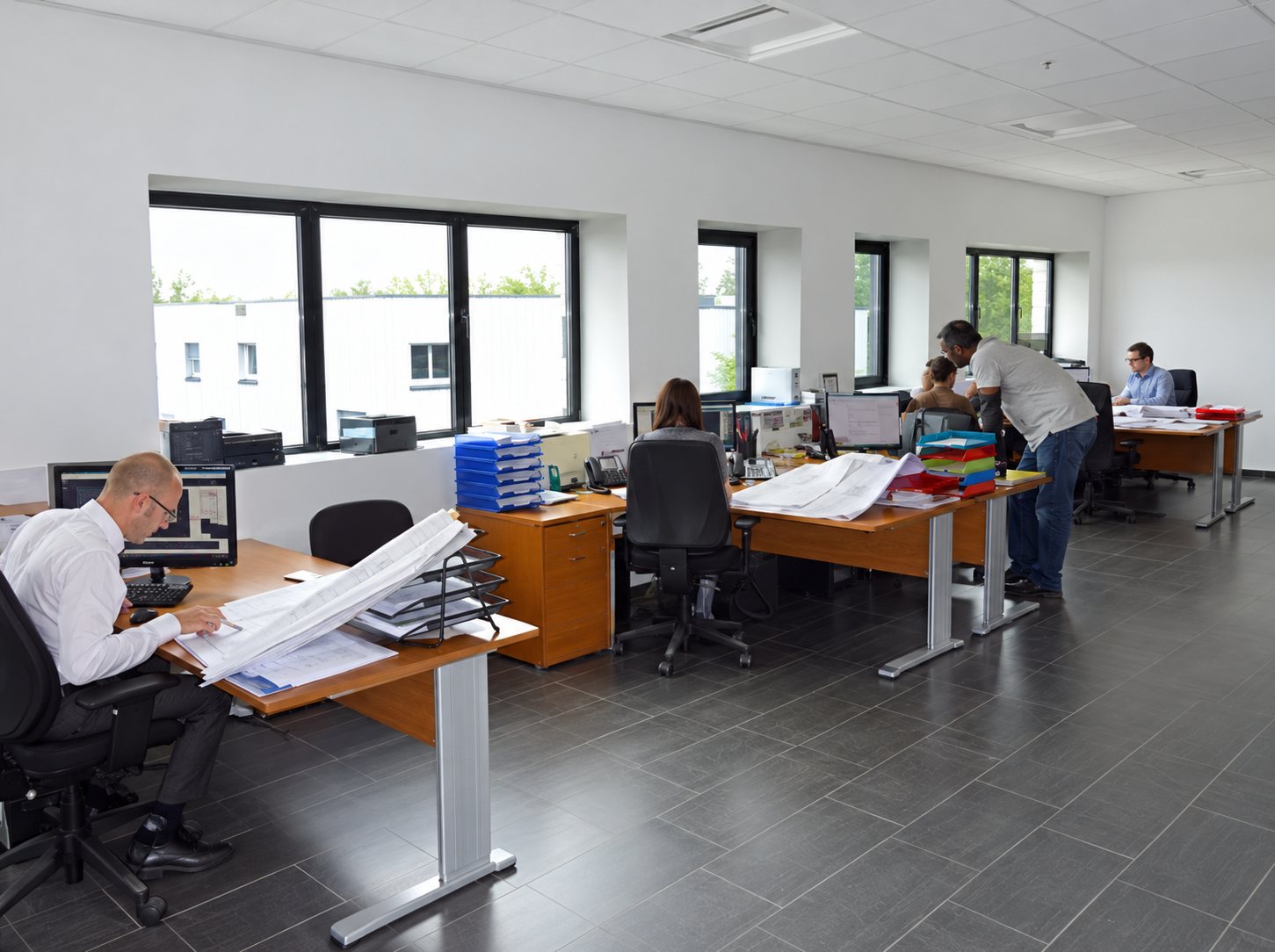 Des employés travaillent à leurs bureaux en bois dans un grand open space lumineux aux larges fenêtres.