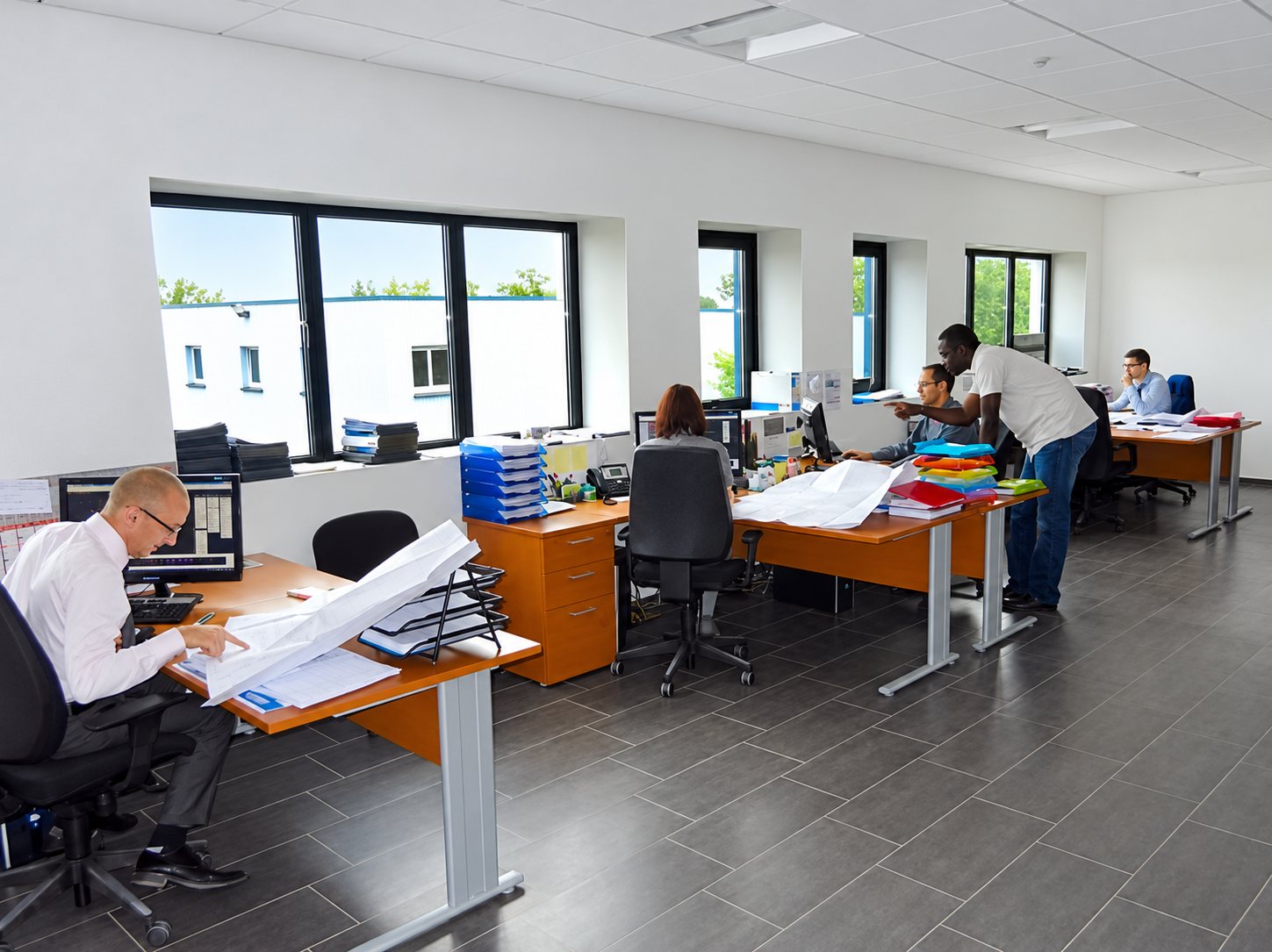 Plusieurs employés travaillent à leurs bureaux en bois dans un grand open space lumineux, certains consultant des plans papier.
