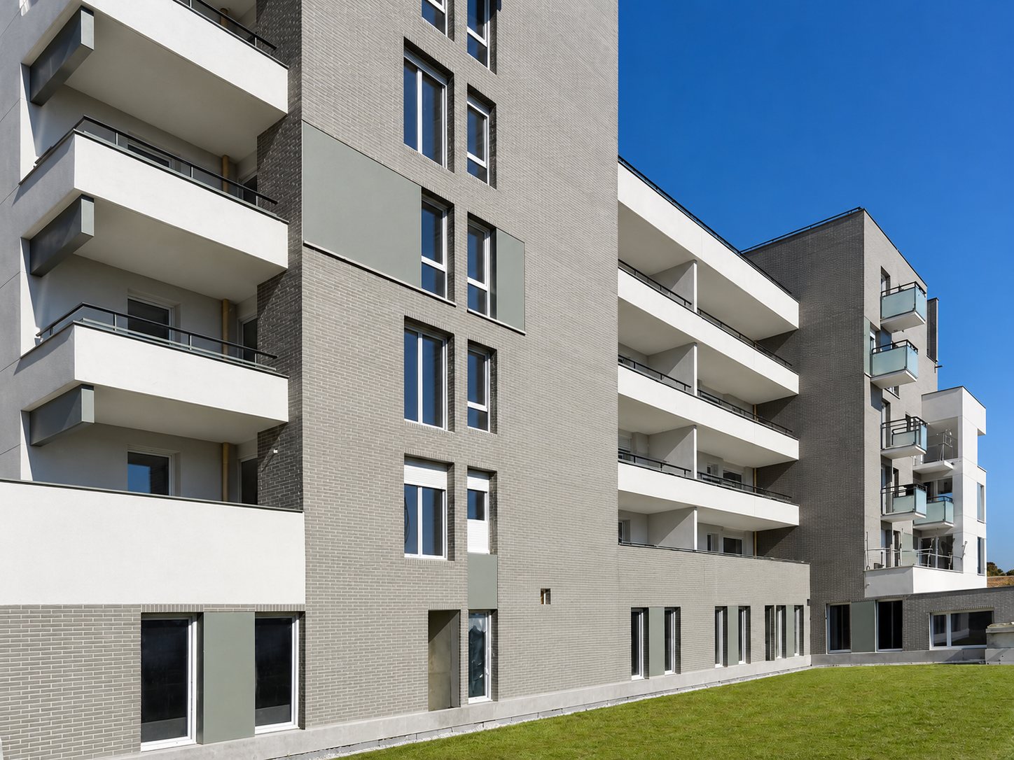 Immeubles résidentiels neufs aux façades grises et blanches avec balcons, au bord d'une pelouse sous un ciel bleu.