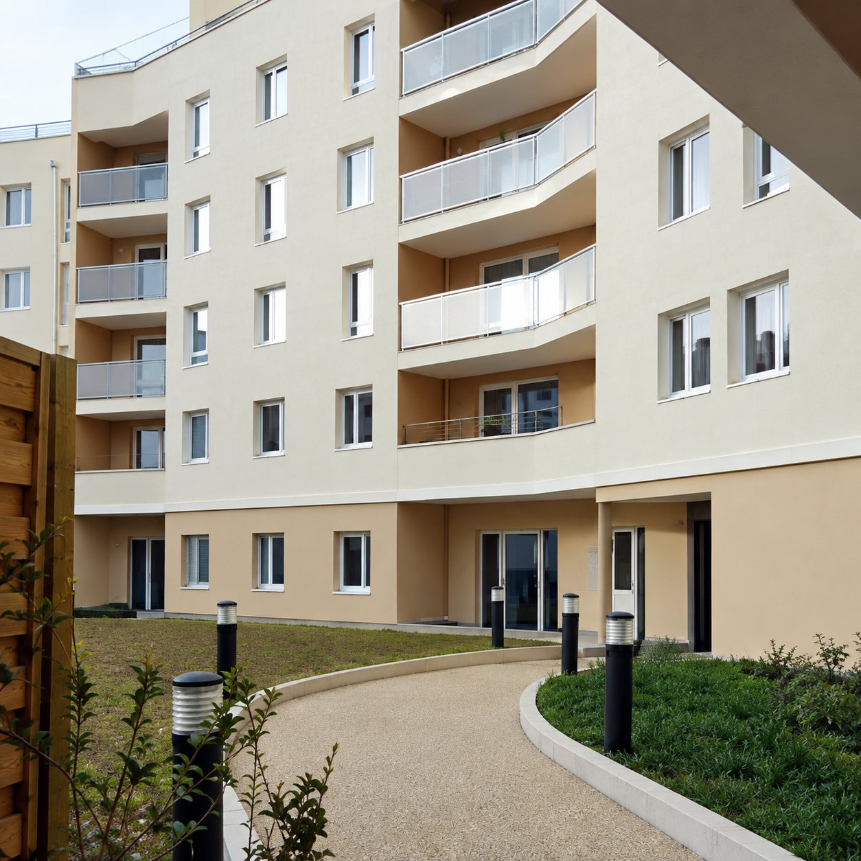 Façade claire d'un immeuble résidentiel récent avec balcons, longée par une allée piétonne et des espaces verts.
