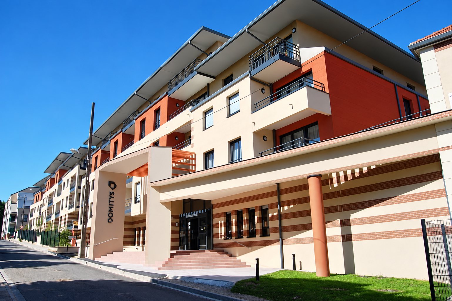 Façade d'une résidence récente aux balcons et aux murs beige et orange, avec l'enseigne Domitys à l'entrée, sous un ciel bleu.