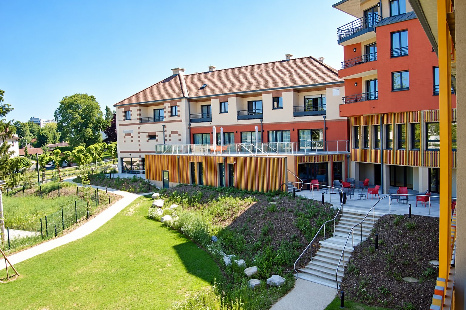 Un bâtiment résidentiel moderne aux façades orange et bois, avec terrasses, mobilier rouge, escalier et jardin paysager au premier plan.