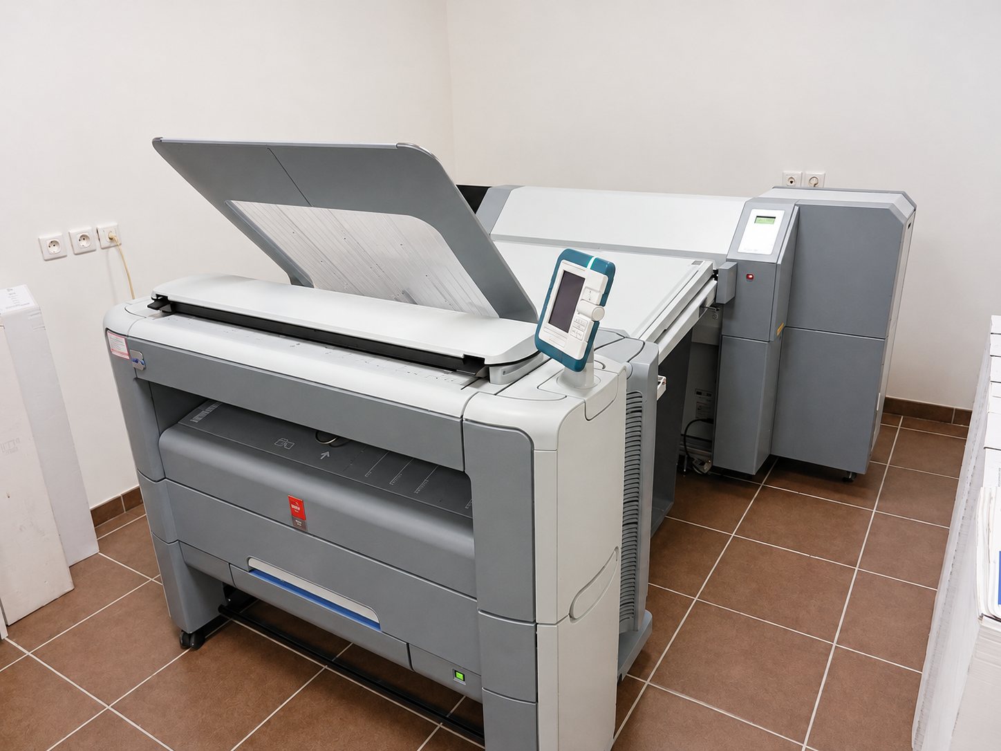 Un traceur grand format gris avec scanner et écran tactile est installé dans une pièce aux murs clairs et au carrelage brun.