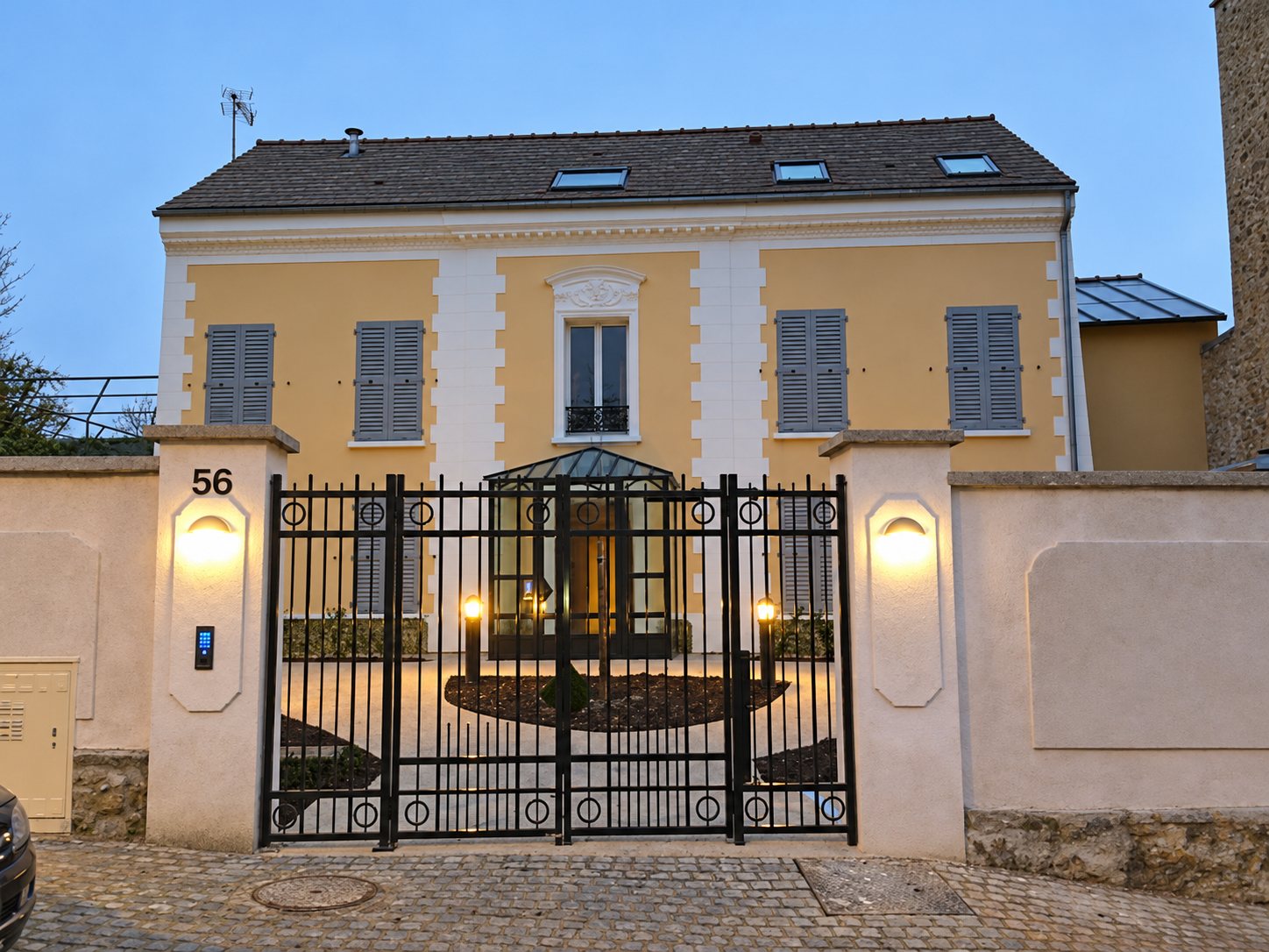 Maison aux façades jaune clair derrière un portail noir éclairé, au crépuscule.