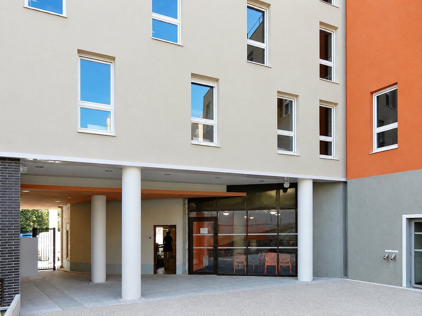 Entrée vitrée d'un immeuble aux façades grises et orange, soutenue par des colonnes blanches.