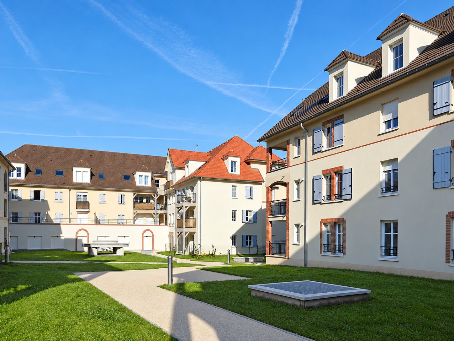 Bâtiments de résidence aux toits d'ardoise et de tuiles, avec pelouse et chemin sous un ciel bleu.